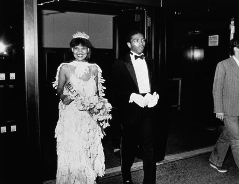 A black-and-white photograph of a smiling Black woman in a beauty pageant gown, crown, gloves, bouquet, and sash is escorted into a room by a young Black man in a tuxedo.