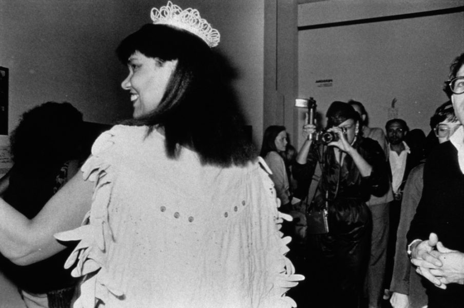 A black-and-white photograph of a Black woman in a beauty pageant gown, crown, gloves, bouquet, and sash is shown in profile as she smiles in profile at someone to her left, while a photographer takes her photo. Her white cape appears made from gloves.