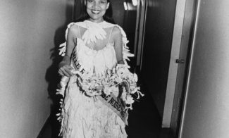 A black-and-white photograph of a Black woman in a beauty pageant gown, crown, gloves, bouquet, and sash  walks down a hallway toward the viewer, smiling widely.