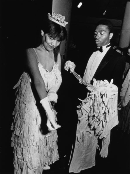 A black-and-white photograph of a Black woman in a beauty pageant gown and crown adjusting her gloves as a young Black man in a tuxedo holds her coat, bouquet, and sash.