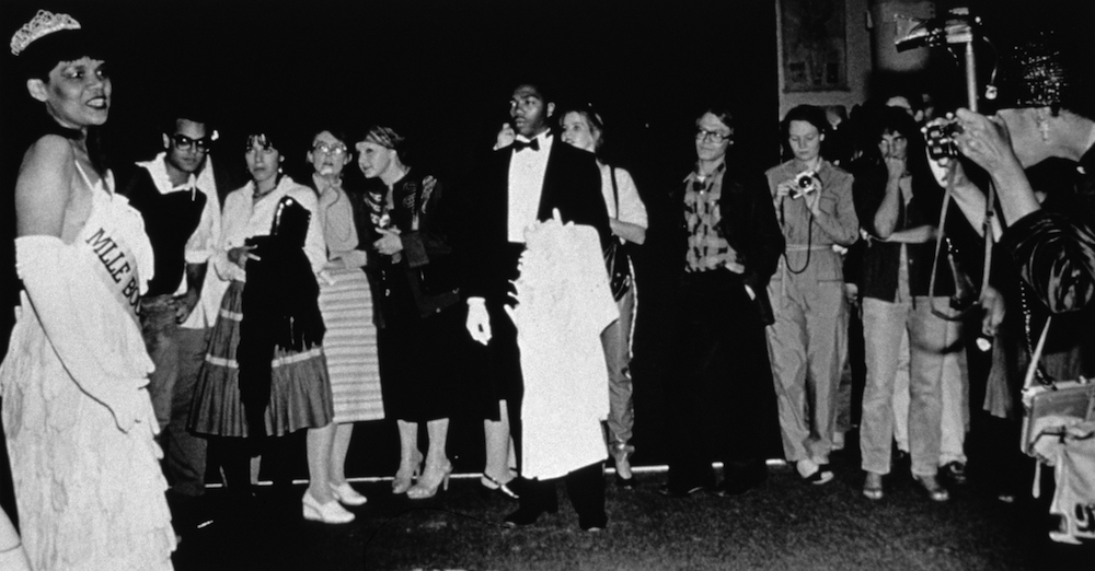 A black-and-white photograph shows a Black woman in a beauty pageant gown, crown, and gloves at the left being watched by a line of less formally dressed people. A young Black man in a tuxedo stands at attention in the center of the frame, holding a white coat, and a photographer shoots the scene from the far right.