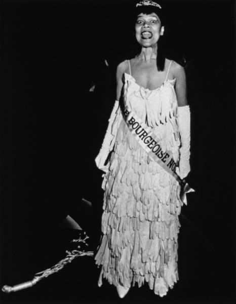 A black-and-white photograph of a Black woman in a beauty pageant gown, crown, gloves, and sash standing in a dark gallery room facing the viewer with her mouth wide open in mid-speech.