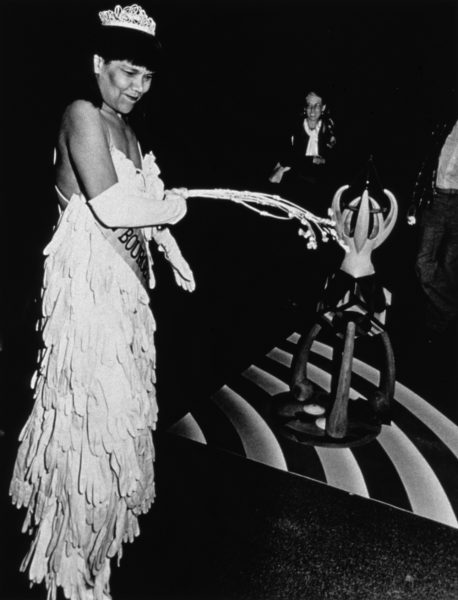 A black-and-white photograph of a Black woman in a beauty pageant gown, crown, and gloves whipping a white cat o’ nine tails in a dark gallery.