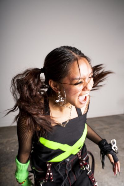 A young woman with black pigtails and medium skin wearing a black top with a large bright green X, gloves, and chains is captured mid-scream.