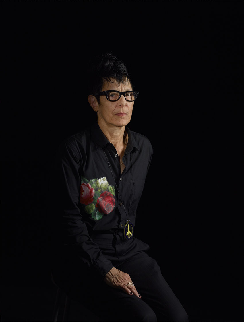 A color photograph shows the choreographer Elizabeth Streb, a pale, middle-aged woman with spiky dark hair, dark-rimmed glasses, black clothing with a large embroidered floral design on the chest, and a serious expression, seated in a three-quarter profile against a black background.