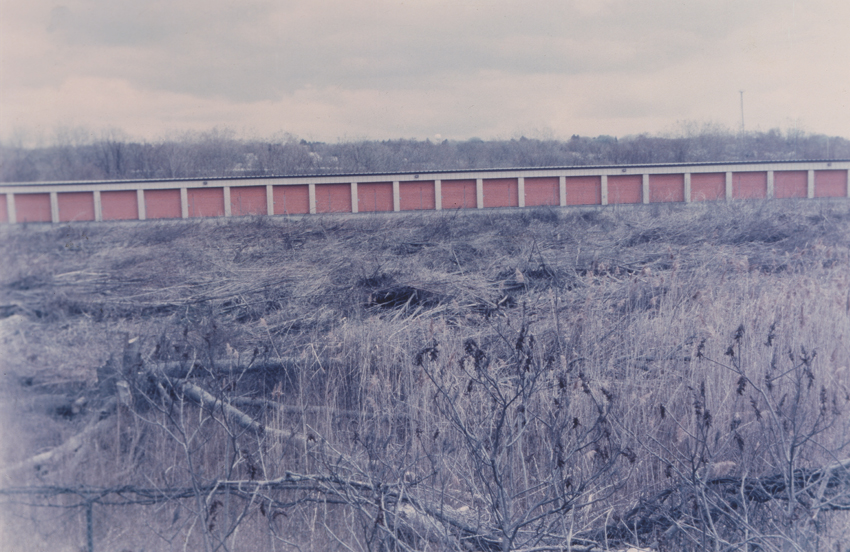 A color photograph depicts a single-story storage facility with a row of orange doors at a distance amid an overgrown, weedy field, rendered in soft purples, blues, and grays.