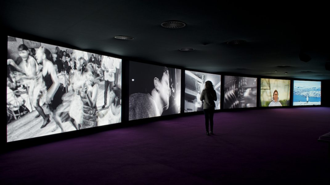 Six large video screens arranged side by side in a gentle curve fill a dark room. A woman stands before them, watching. They each display a different image, some black and white, including people dancing, some color. 