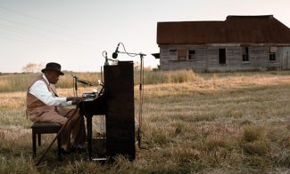 A video still shows musician Pinetop Perkins, an older Black man, playing an upright piano in an open field before a wooden house.