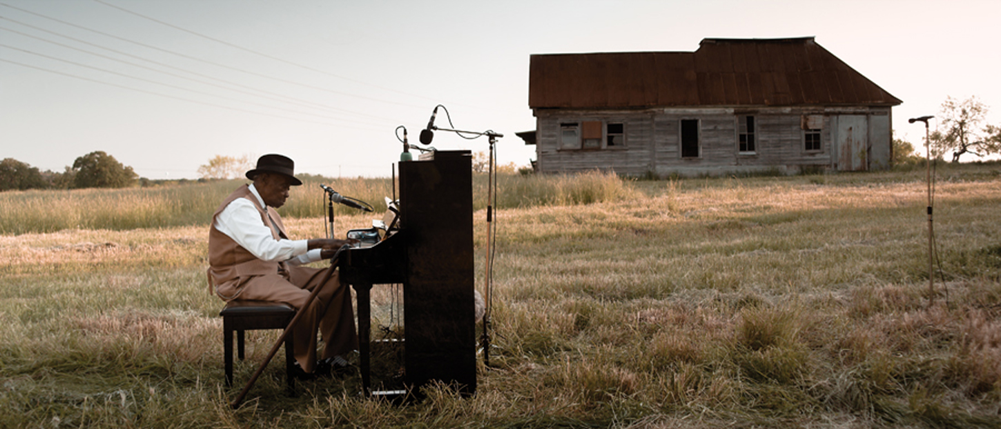 A video still shows musician Pinetop Perkins, an older Black man, playing an upright piano in an open field before a wooden house.