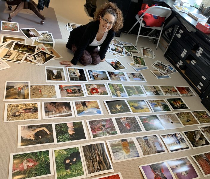 A woman kneels in an art studio amid dozens of photographs laid out on the floor. She is wearing yoga pants, a white shirt, and a cardigan, and smiling at the camera.