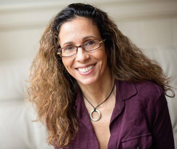 A middle-aged woman with long, wavy, gold-highlighted hair, copper-colored glasses, and a smile. A purple button-up shirt open at the neck frames a heavy silver ring on a dark string around her neck. 