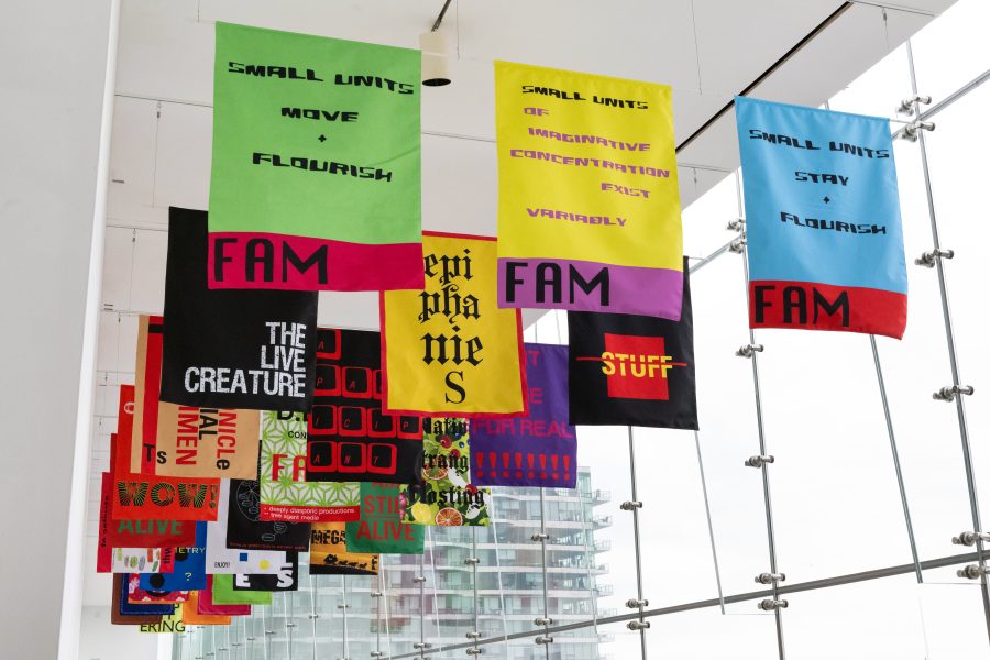 Brightly colored printed flags installed on the ceiling in rows in a white gallery with glass windows on the right