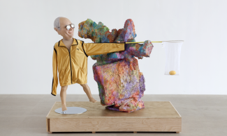 A mixed-media sculpture of a figure built from a Dick Cheney face mask, sunglasses, and a gold jacket along with an amorphous, colorful sculpture on a wooden plinth