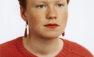 A color photograph portrait of a young woman with red hair, pale skin, red lipstick, and an orange sweater, shown in three-quarter profile.