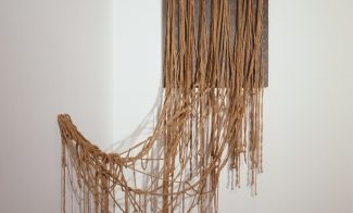 An installation of ochre- colored string attached in a regular pattern to a piece of grey painted plywood but tangling as they fall to the floor or are attached loosely to the adjacent wall.