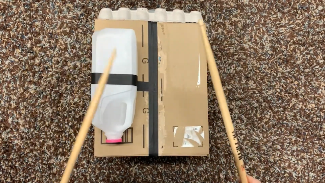 An aerial image of a pair of hands holding sticks while striking a DIY drum made of a box, a milk carton, and an egg carton.
