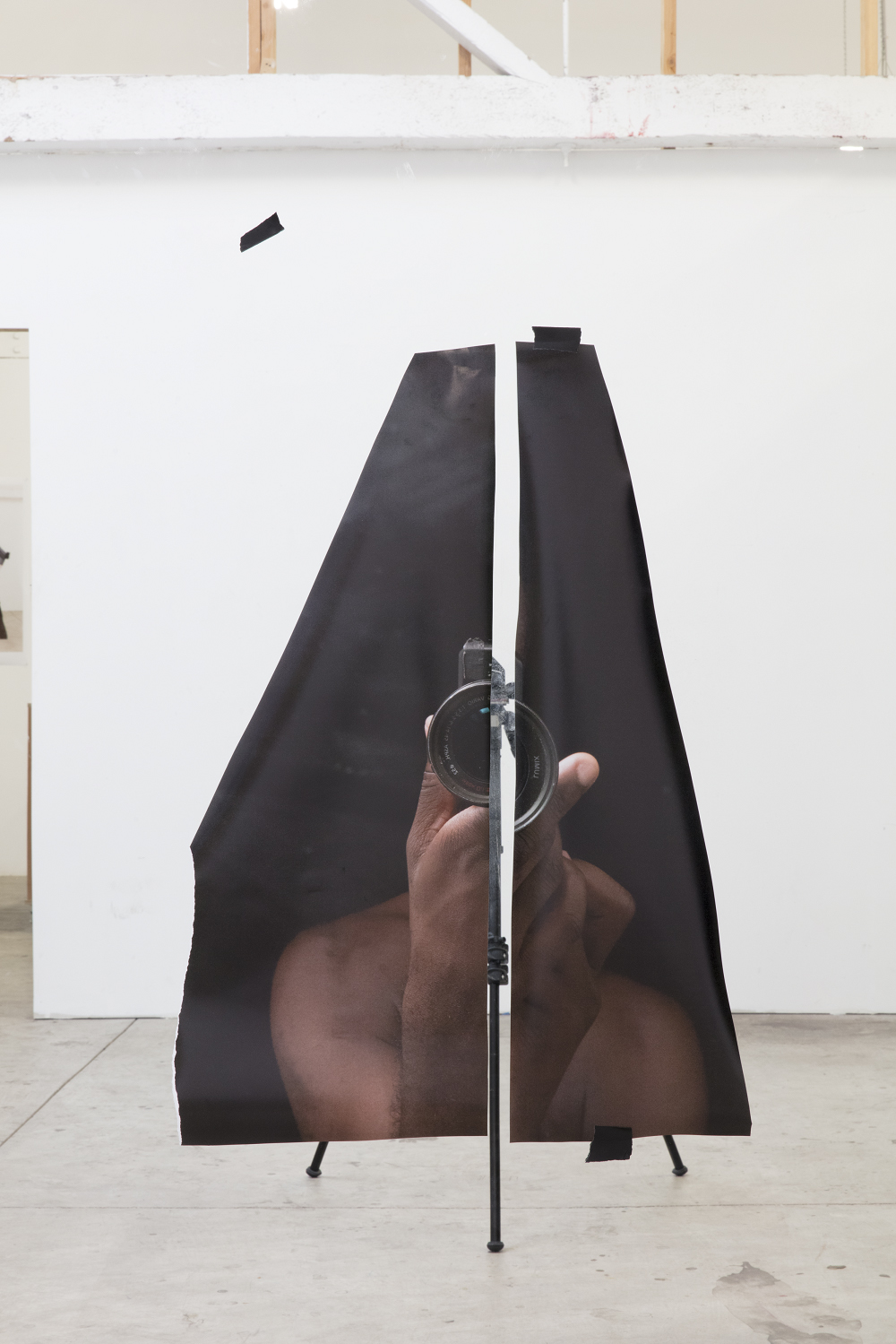 A color photograph shows a large photograph, cut vertically in two, apparently floating in a white-walled studio. The cut image depicts the shoulders and hands of a dark-skinned man, holding a camera. In the space between the two pieces a camera tripod can be seen.