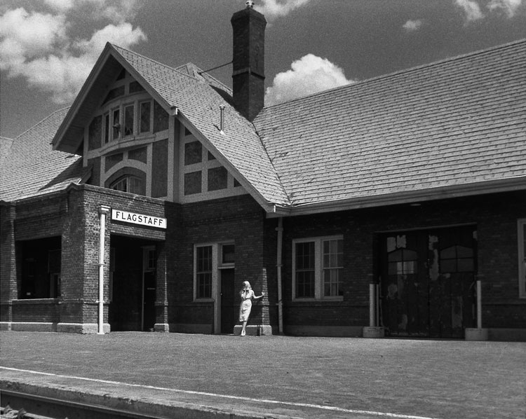 A black-and-white photograph of the artist, a white woman, posed on the empty outdoor platform of the Flagstaff train station and captured from across the tracks under a partly cloudy sky.