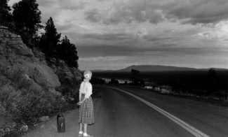 A black-and-white photograph of the artist with blond hair, a white blouse, and a calf-length plaid skirt standing on the side of an open road in a hilly evening landscape, her back turned to the viewer and a suitcase by her side on the ground.
