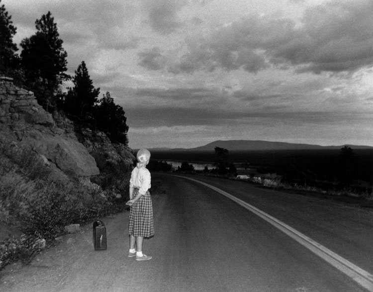 A black-and-white photograph of the artist with blond hair, a white blouse, and a calf-length plaid skirt standing on the side of an open road in a hilly evening landscape, her back turned to the viewer and a suitcase by her side on the ground.
