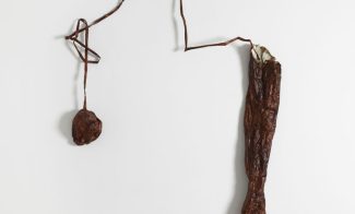 A dark red wall sculpture of a lumpy ball form connected by a coiled and looped string to a disembodied forearm and hand.