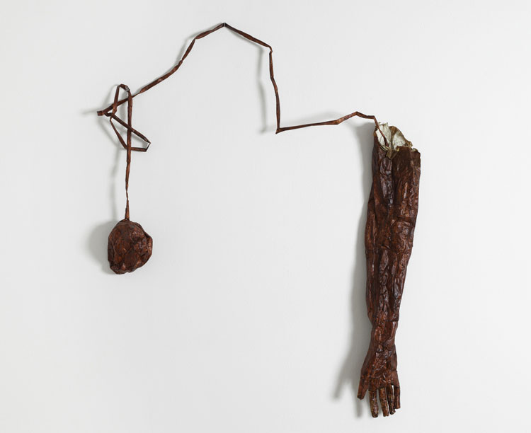 A dark red wall sculpture of a lumpy ball form connected by a coiled and looped string to a disembodied forearm and hand.