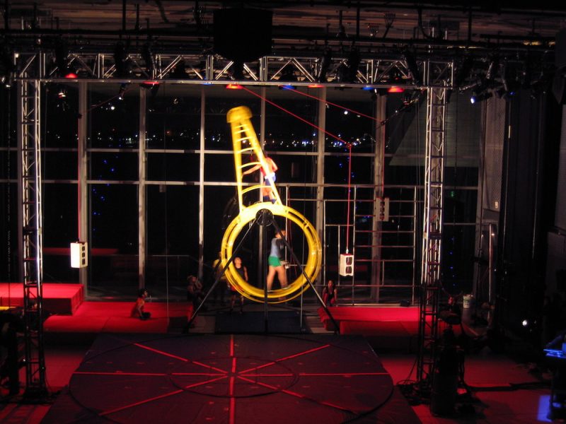 A slightly blurry photo shows several dancers inside and on top of a yellow human-sized hamster wheel on a stage full of scaffolding.