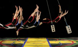 Five dancers seem to fly several feet off a colorful stage floor while two cinderblocks suspended on wires swing among them.