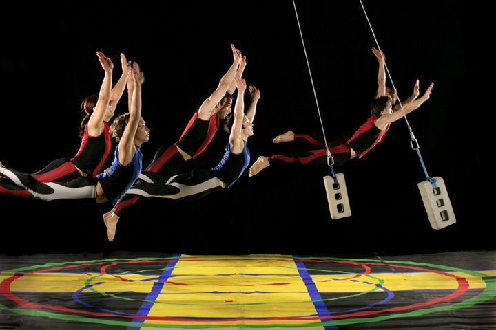 Five dancers seem to fly several feet off a colorful stage floor while two cinderblocks suspended on wires swing among them. 