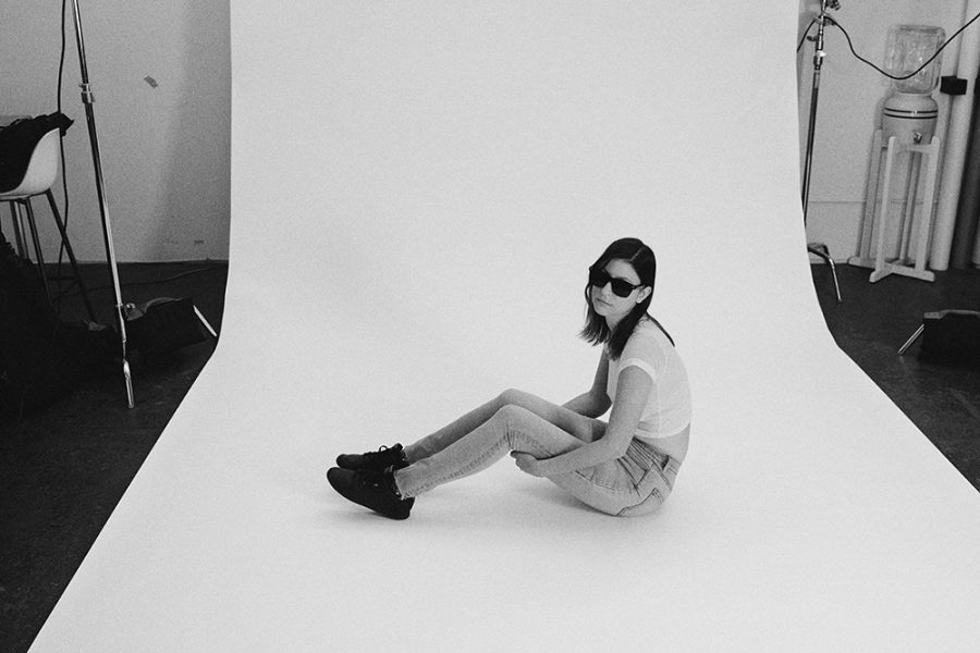 A black and white photo of someone with shoulder length hair and sunglasses sitting on a white backdrop