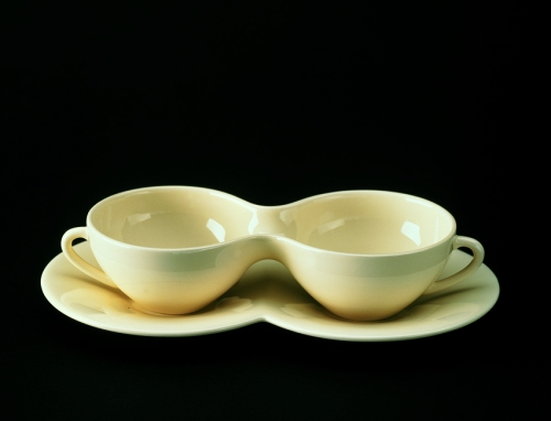 A sculpture of two glazed white teacups on matching saucers. The cups are fused together to appear conjoined, as are the saucers.