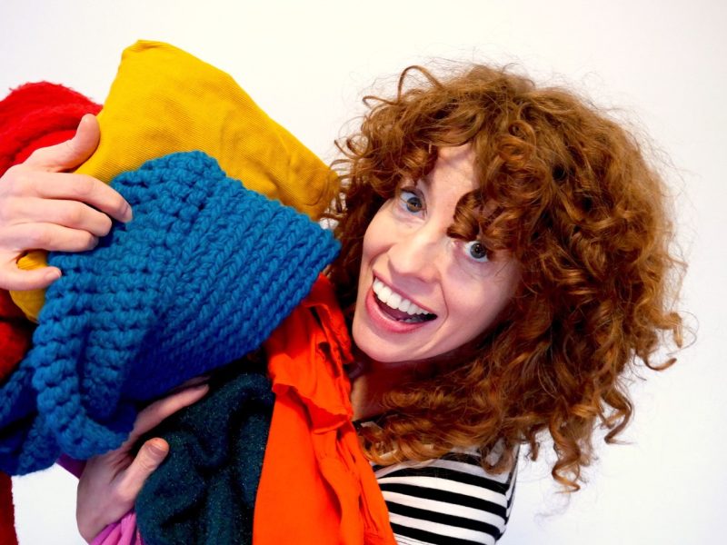 A woman with curly red hair smiling at the camera with piles of knits and fabrics in her arms.