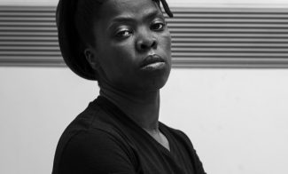 A portrait of artist Zanele Muholi with their head raised and arms crossed looking into the camera.