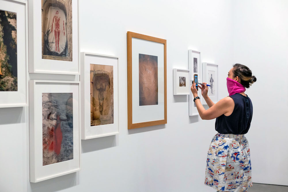 A young woman in a face covering takes a cell phone photo of a wall of artworks by Ana Mendiata, Mona Hatoum, and others in the exhibition 