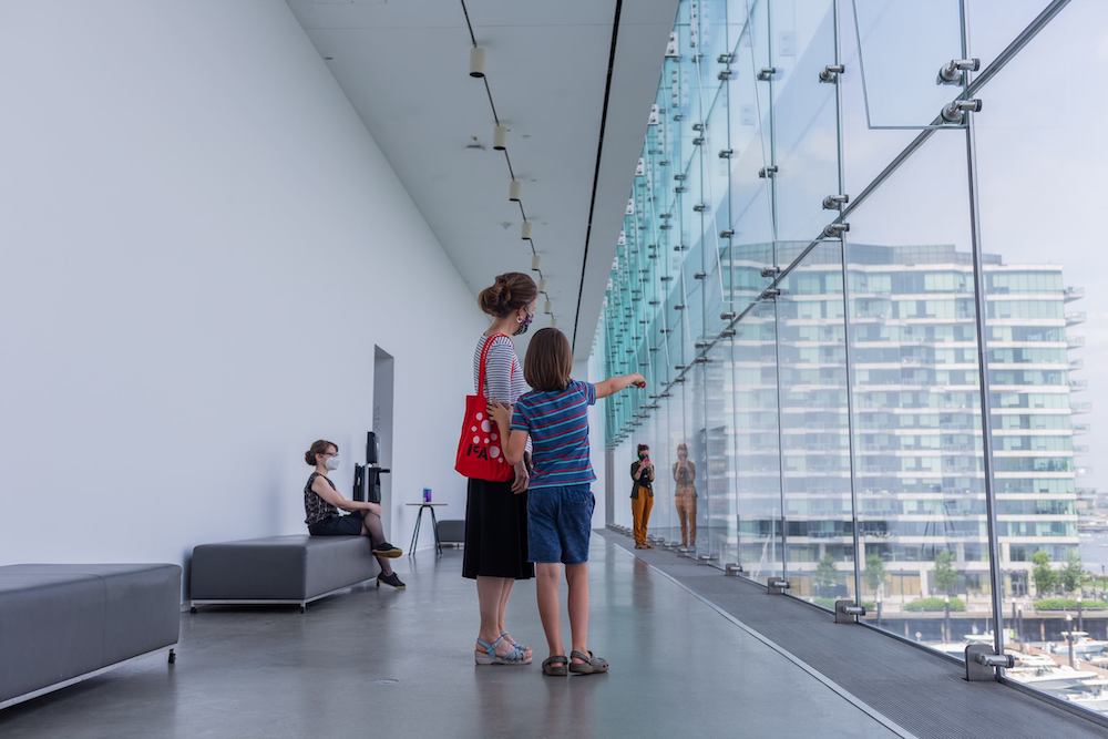 A school-age child and his mother look out of a wall of windows in the ICA's Founders Gallery. The boy points.
