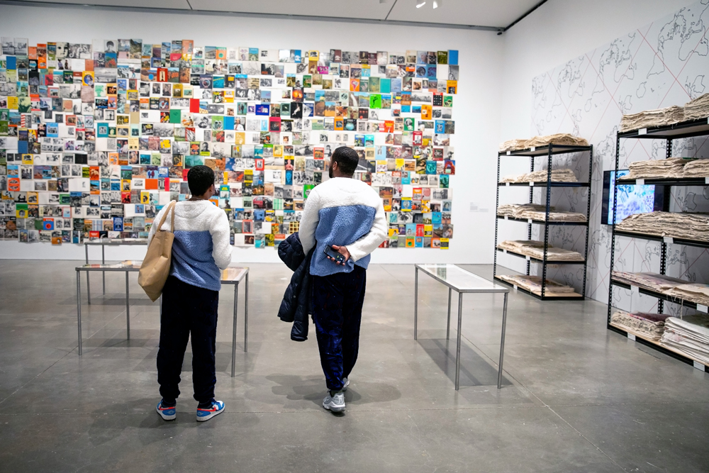 Two people in matching outfits look at wall of book installation while surrounded by glass desk cases