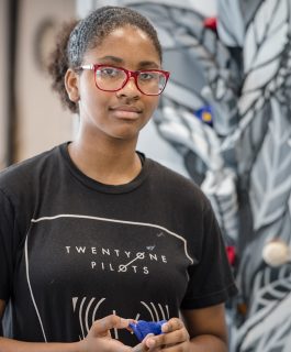 A headshot of Amaya Willis wearing a black t-shirt in front of a black and white artwork depicting a leaf.