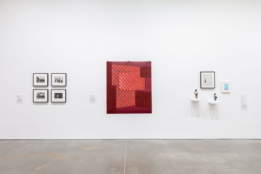 A wall with a red, unframed painting in the middle showing a lighter red woven mesh. To the left, four framed black and white photos hang in a grid. On the right, two small sculpted figures sit on short shelves. Two small framed works hang above them.