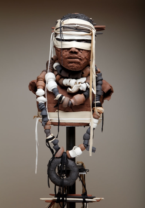 A brown clay bust on a pole with white cloth covering its eyes and clay chains around its neck