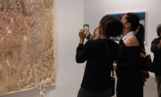 Two visitors look at a hanging brown painting and take a picture of it