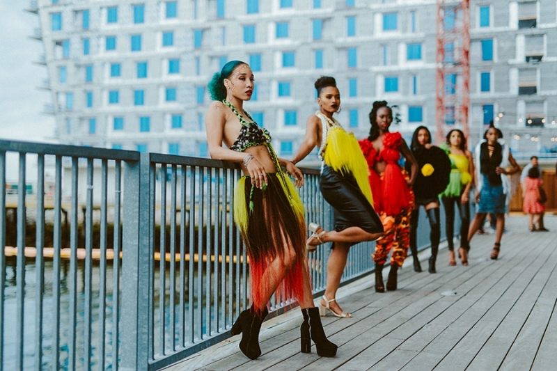 Six models with medium to dark skin pose in colorful clothing along a railing overlooking Boston Harbor behind the ICA during a Caribbean Fashion event. 