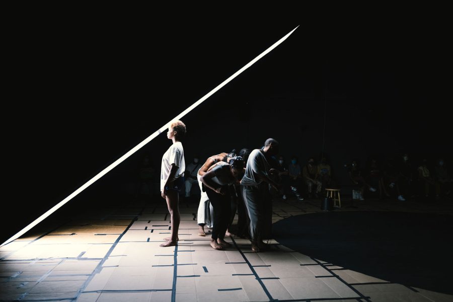 A performer faces a beam of light while other performers are behind them in the darkness