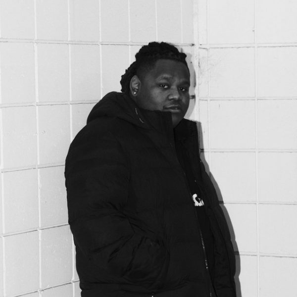 A person wearing a dark puffer jacket stands against a tiled wall, looking at the camera with a neutral expression. Captured in black and white, the photo echoes the cool tone of Harborwalk Sounds at Berklee.