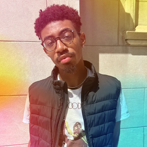 A young man with glasses and short curly hair stands against a stone wall, wearing a black puffer vest over a printed T-shirt. Soft rainbow light effects evoke the creative energy of Berklee's Harborwalk Sounds across the photo.