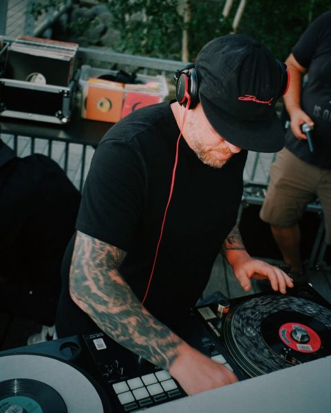 A tattooed DJ wearing a black t-shirt, black cap, and headphones mixes vinyl records at an outdoor Vinyl Nights DJ event at ICA Boston, with equipment and records visible around him.