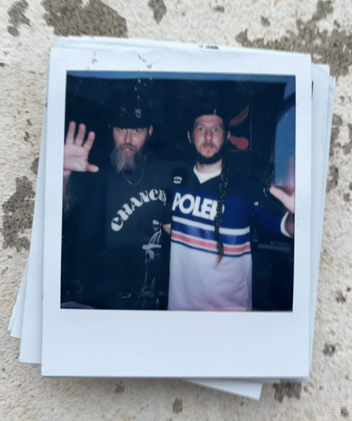 Two men pose for a photo at ICA Boston, both raising one hand toward the camera. One has a long beard with a hat; the other sports a long braid and POLO jersey. The photo, taken during Vinyl Nights DJ, rests on a textured surface.