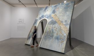 A blurry figure walks out of a blue sculptural arch in a gallery
