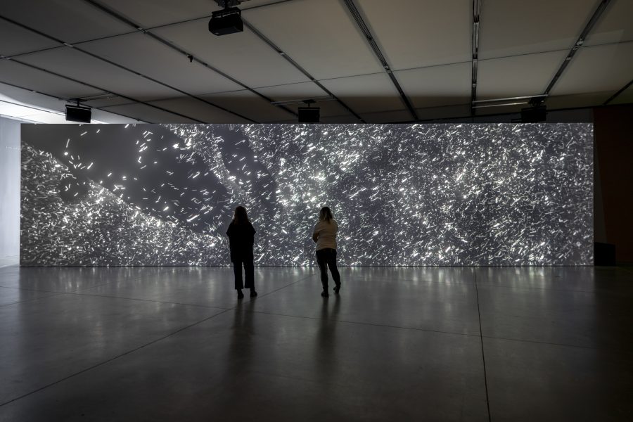 Two people in dark gallery looking at wide floor to ceiling video screen of white flecks against dark background