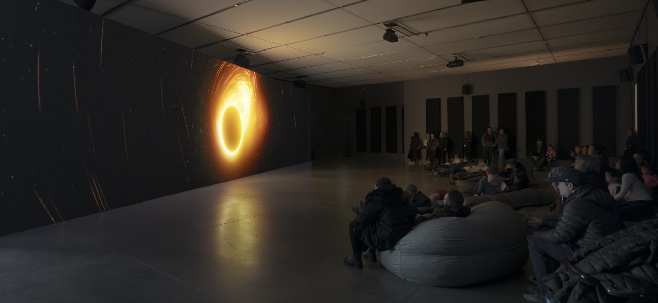 People in dark gallery seated on beanbags and standing, watching a large video installation with orange fiery circle