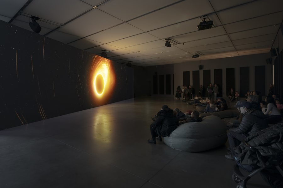 People in dark gallery seated on beanbags and standing, watching a large video installation with orange fiery circle